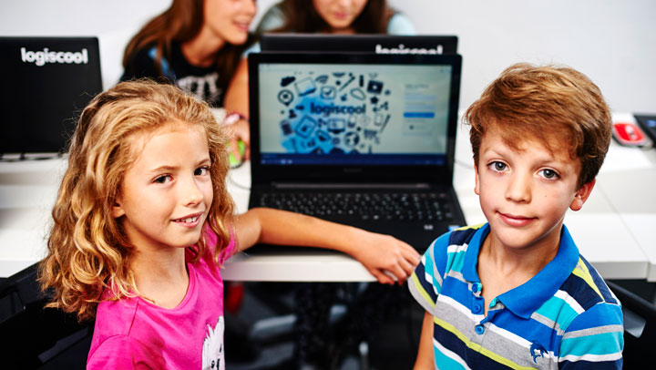 blond meisje in roze t-shirt en blonde jongen in blauw gestreept t-shirt kijken naar de camera met een laptop op de achtergrond